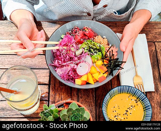 Woman eating tasty colorful healthy natural organic vegetarian Hawaiian poke bowl using asian chopsticks on rustic wooden table