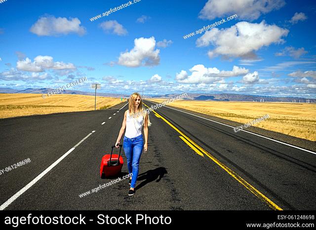 Attractive blond woman pulling her red luggage and walking away from it all on an empty countryside road