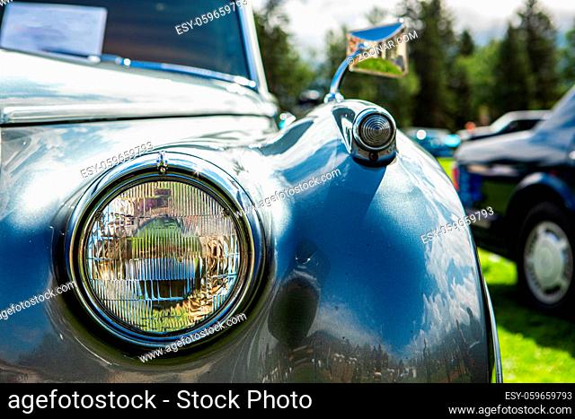 bright metal grey old classic antique American car front part, right side, close up glass headlight with chrome frame, blurred cars outdoor background