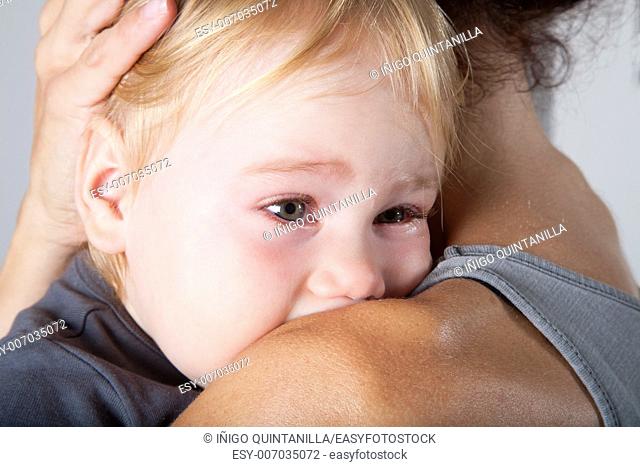 portrait of one year age blonde lovely cute caucasian white baby grey shirt looking face crying and scream shout with tears eyes in brunette woman mother arms...