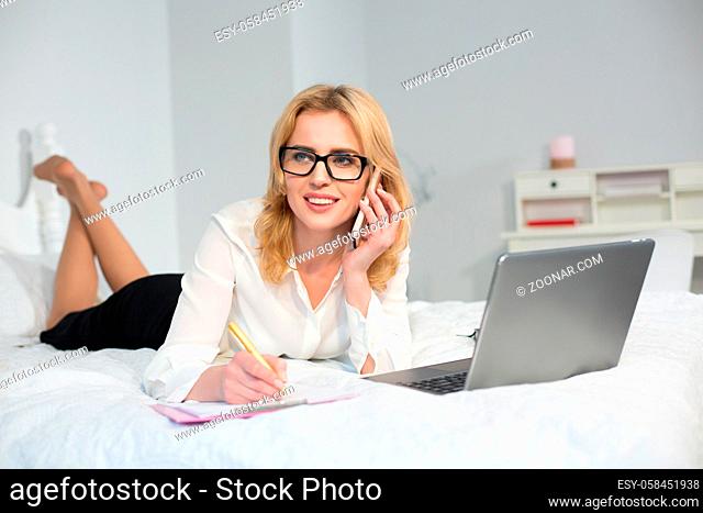 Beautiful businesswoman making business in bed. Speaking over phone and working on computer