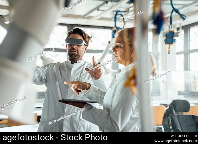 Senior scientist using laptop with colleague wearing virtual reality simulator in laboratory