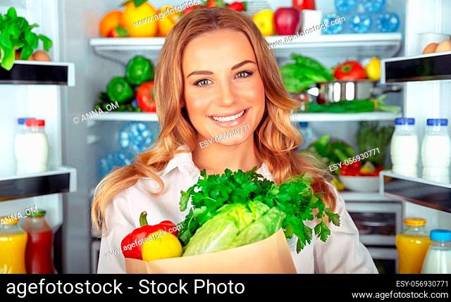 Vegetarian lifestyle, portrait of a beautiful woman standing near open fridge full of different fresh vegetables, organic nutrition, weight control