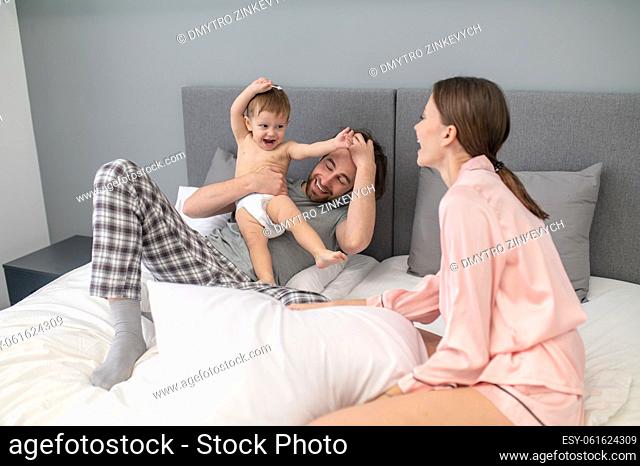 Happiness. Happy young woman sitting looking at playful little cute baby in mans arms lying on bed in bedroom