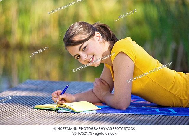 Teenaged girl lying on a footbridge and writing in her diary, smiling at the camera