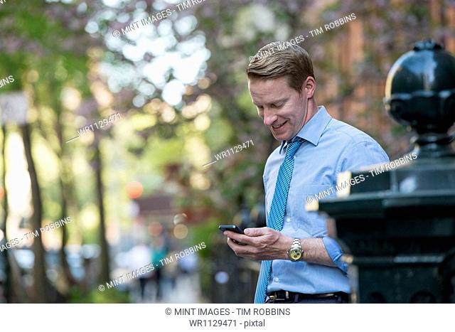 A man in a business shirt and tie outdoors in a city park. Looking down and checking his phone