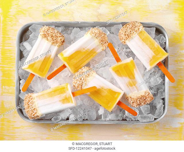Mango and coconut ice lollies topped with white chocolate and desiccated coconut on a bed of ice cubes