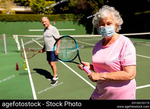 Portrait of senior caucasian couple wearing face mask looking at camera and smiling on tennis court