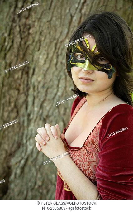 Young woman, standing by a tree in a park, wearing a mask and red vintage dress
