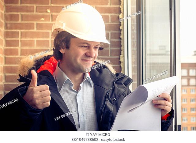 Handsome architect is standing near building outdoors. He is giving thumb up and smiling. The man in helmet is looking at camera happily