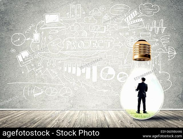 Businessman inside of light bulb in empty concrete room and sketches on wall
