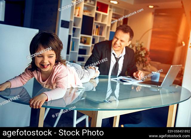 Little daughter lying on table and smiling while her father working on laptop. Business at home concept