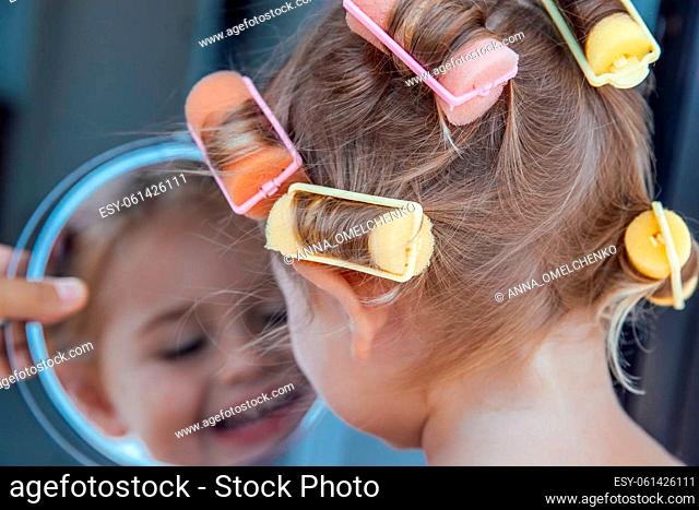 Portrait of a Nice Little Baby Girl with Colorful Curlers on Hair Enjoying her Reflection in the Mirror. Happy Childs Fun. Active Childhood