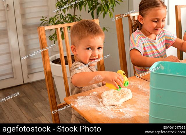 The cute baby is sitting at a table in the kitchen and playing with a toy. The boy is playing while cooking pizza