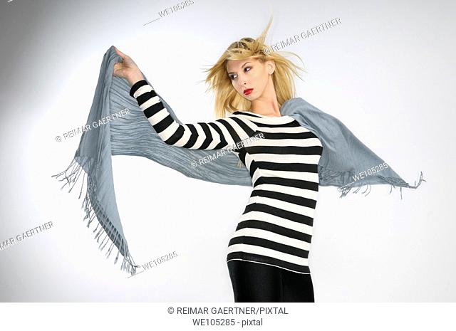 Young blond woman in striped top holding a scarf in the wind on gray background
