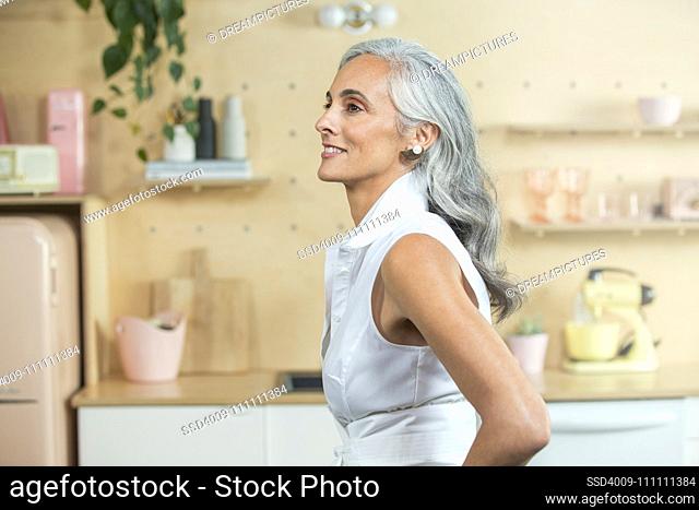 Standing in a modern kitchen, A youthful middle-aged woman with gray hair looks off camera