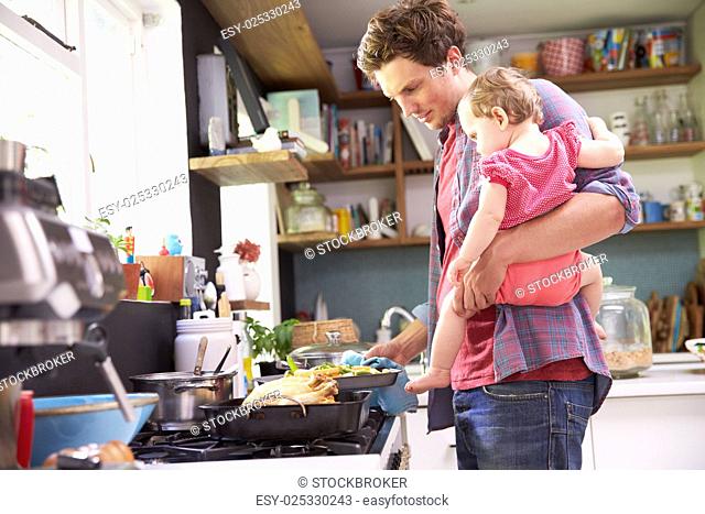 Father Cooking Meal Whilst Holding Daughter In Kitchen