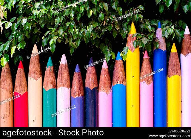 Unusual bright colour fence made of tall big pencils in row, red, blue, yellow, white, green colors, multi colored wall and green tree foliage outdoors