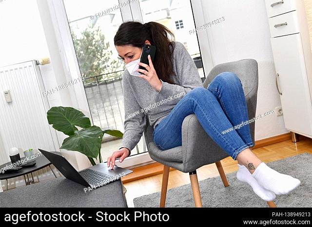 A young woman is sitting in her apartment with a laptop and is working from home because of the corona pandemic, wearing an FFP2 mask and holding a smartphone