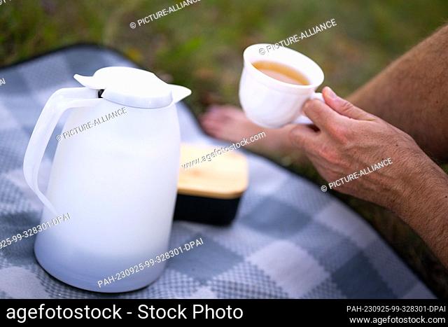 ILLUSTRATION - 17 September 2023, Berlin: A thermos flask stands on a picnic blanket, while in the background a man holds a cup of warm tea in his hand