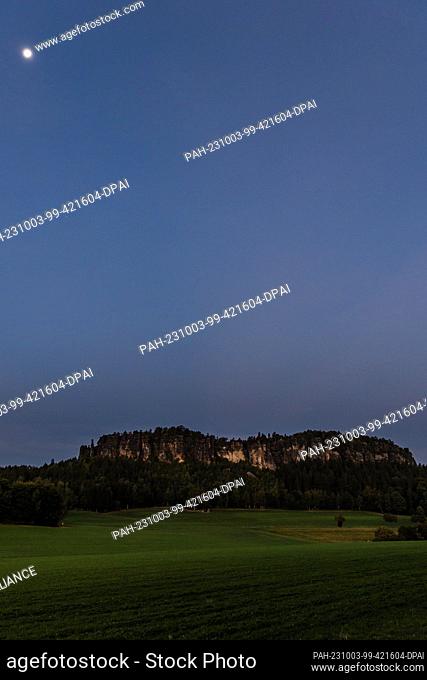 03 October 2023, Saxony, Pfaffendorf: Shortly before sunrise the moon is above the Pfaffenstein in Saxon Switzerland on German Unity Day