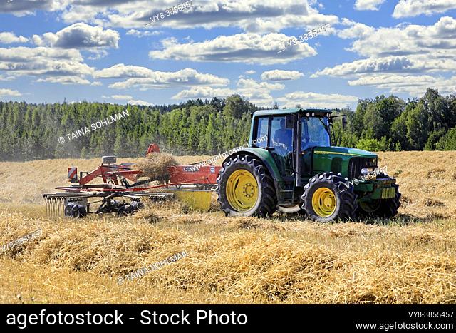 Farmer working on field with John Deere 6420S tractor and Fella rotary rake, raking dry straw on day of late summer. Salo, Finland. August 16, 2020