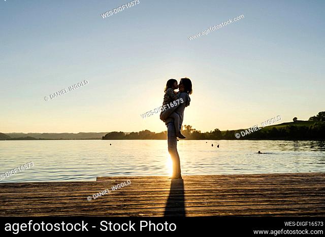 Mother kissing daughter while standing on pier at sunset