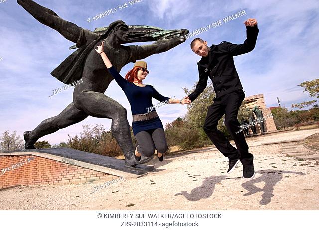 Hungarian man and woman jumping in front of monument while visiting Memento Statue Park in Budapest, Hungary