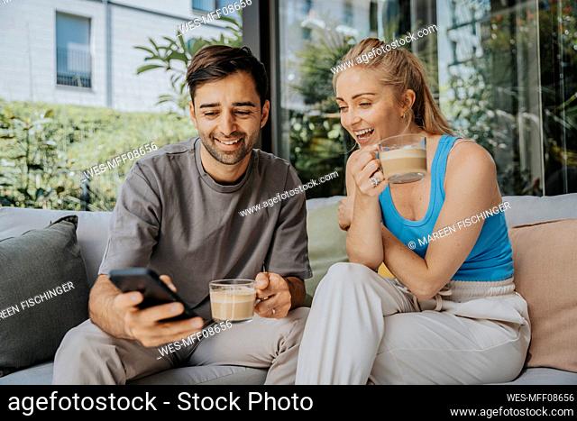 Man sharing smart phone with woman having coffee on sofa in lounge