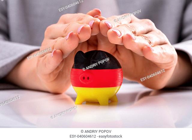 Close-up Of A Woman's Hand Protecting Piggybank Painted With German Flag On Desk