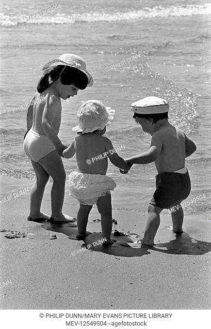 Three small children wearing sun hats hold hands in the sunshine and walk to the sea on the beach at Tenby, Pembrokeshire, Wales
