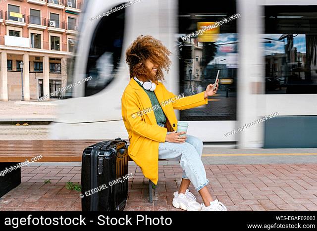 Smiling Afro woman with tousled hair taking selfie through mobile phone while sitting on bench at tram station