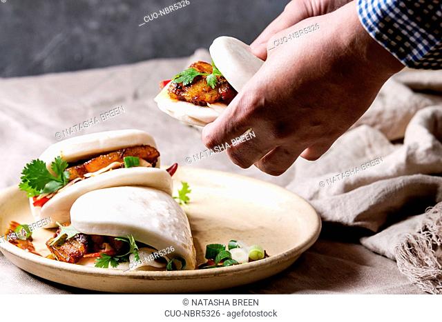 Man's hands hold asian sandwich steamed gua bao buns with pork belly, greens and vegetables served in ceramic plate on table with linen tablecloth