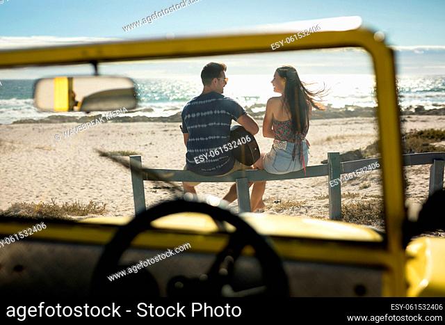 Happy caucasian couple next to beach buggy by the sea playing guitar