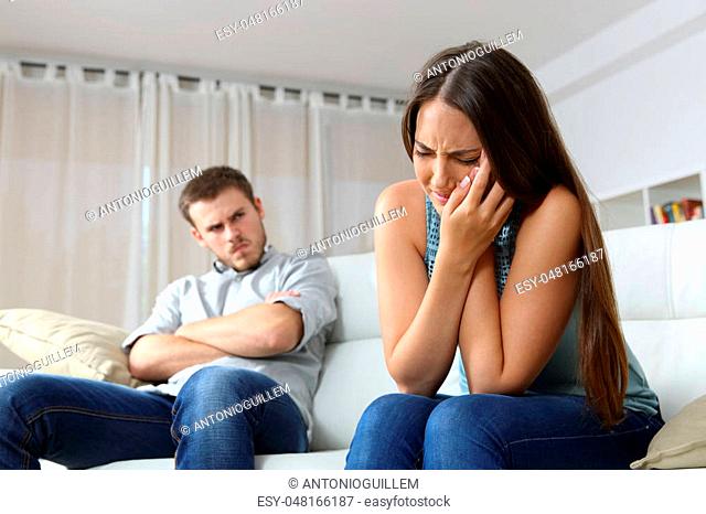 Woman lamenting domestic violence beside her angry husband sitting on a couch in the living room in a house indoor
