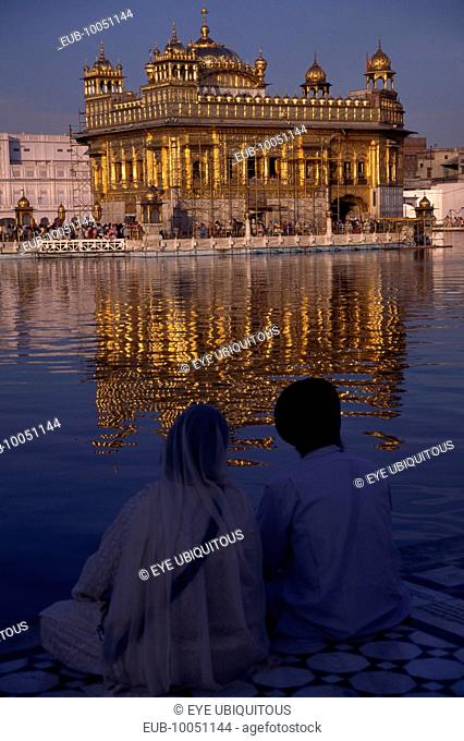 Sikh couple sitting on marble walkway looking out across sacred pool towards the Golden Temple and shimmering reflection on rippled water surface