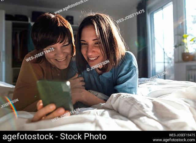 Curious lesbian couple sharing smart phone lying on bed at home