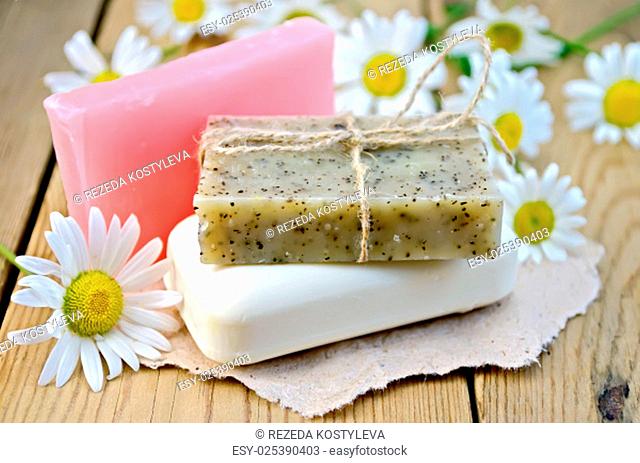 Three bars of soap pink, brown and white on a piece of paper, daisy flowers on a background of wooden boards