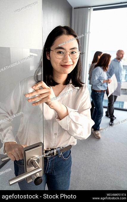 Portrait of young businesswoman opening door with colleagues in background in office