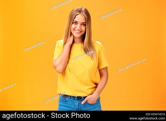 Portrait of shy and timid feminine girl with tan and straight fait hair rubbing neck and smiling sensually with happy carefree expression holding hand in pocket...