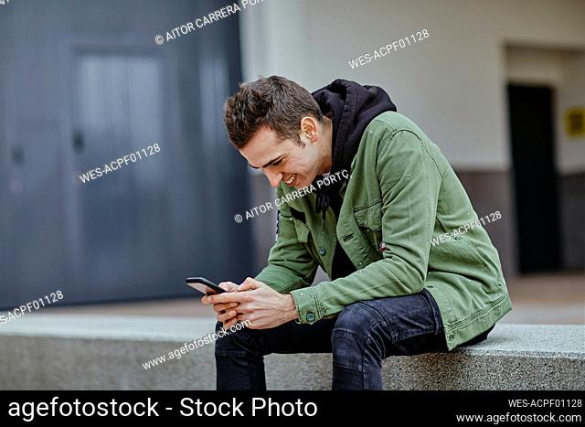 Smiling handsome man using smart phone while sitting on retaining wall at sidewalk cafe