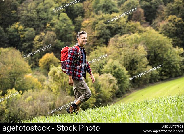 Smiling mature man with backpack walking on grass in front of trees