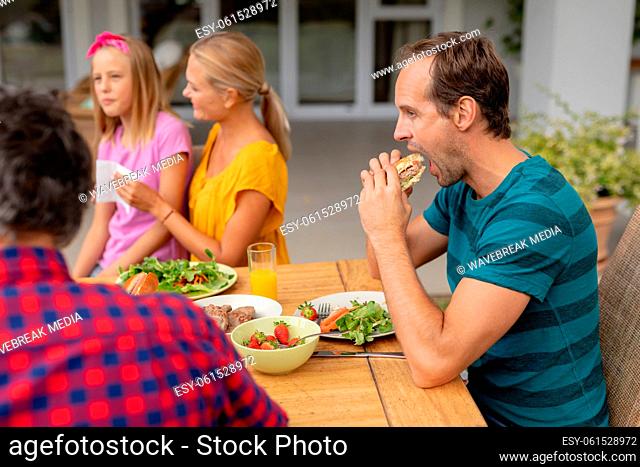 Caucasian man eating hamburger sitting at table with family having meal in garden