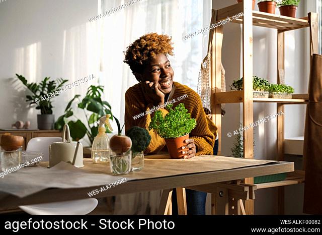 Happy woman with potted plant leaning on table at home