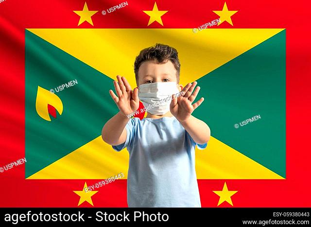 Little white boy in a protective mask on the background of the flag of Grenada. Makes a stop sign with his hands, stay at home Grenada