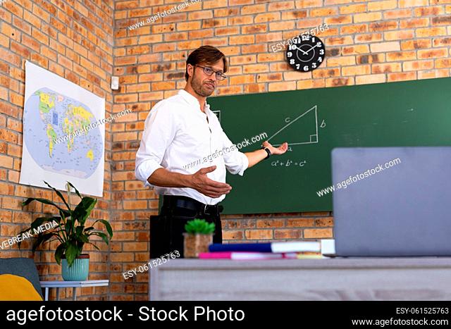 Caucasian male maths teacher standing by blackboard giving online lesson using laptop