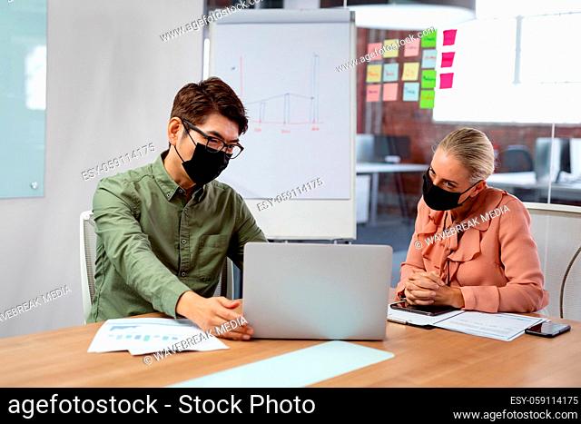 Diverse male and female colleague in face masks sitting at table looking at laptop and discussing