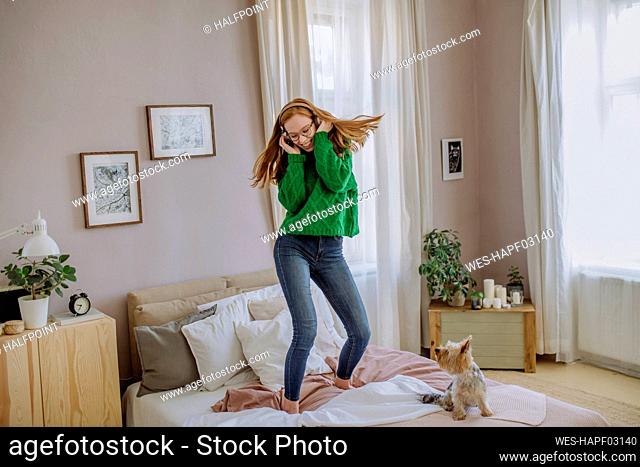 Happy woman listening music through wireless headphones enjoying dance on bed by dog at home