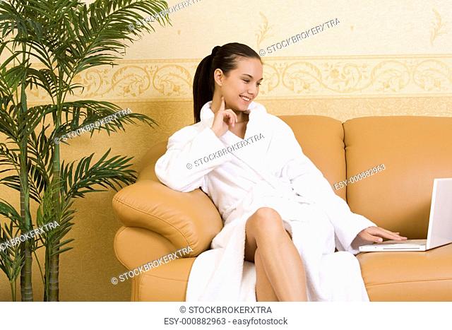 Image of pretty girl in bathrobe looking at laptop monitor while sitting on leather sofa
