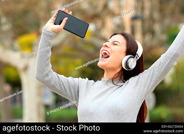 Happy teen singing listening to music showing smart phone screen in a park